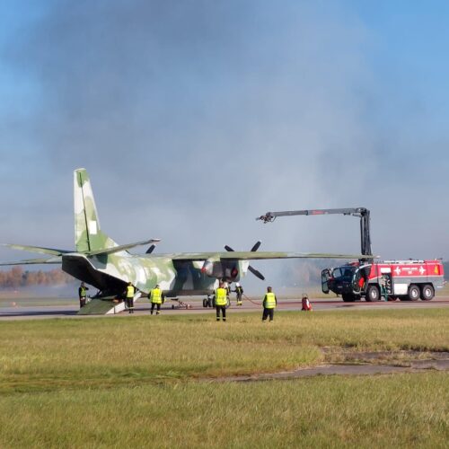 Na trawaiastym terenie lotniska stoi duży samolot wojskowy, który gasi wóz strażacki.