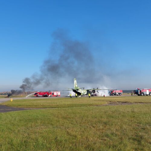 Widok ogólny lotniska. Wozy strażackie biorą udział w akcji gaszenia samolotu. W powietrzu unosi się dużo dymu.